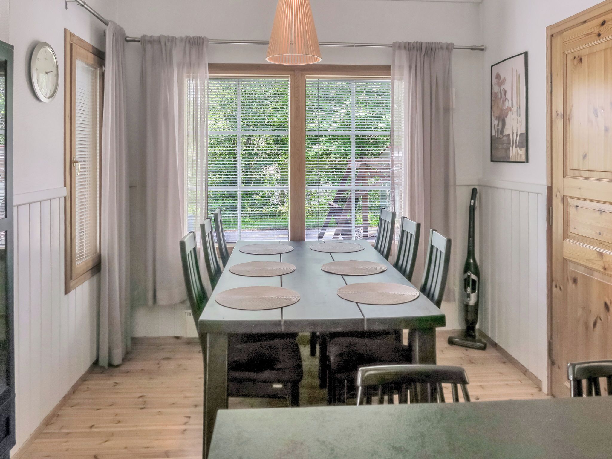 Ein Essbereich mit einem langen Tisch, Stühlen, einer Uhr und einem Fenster mit Jalousien.