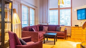 A living room with a sectional sofa, a coffee table, and a leather rug. There are windows with blinds, a television, and a cabinet with glass doors. - Katinkulma 2 by Interhome (Sotkamo)