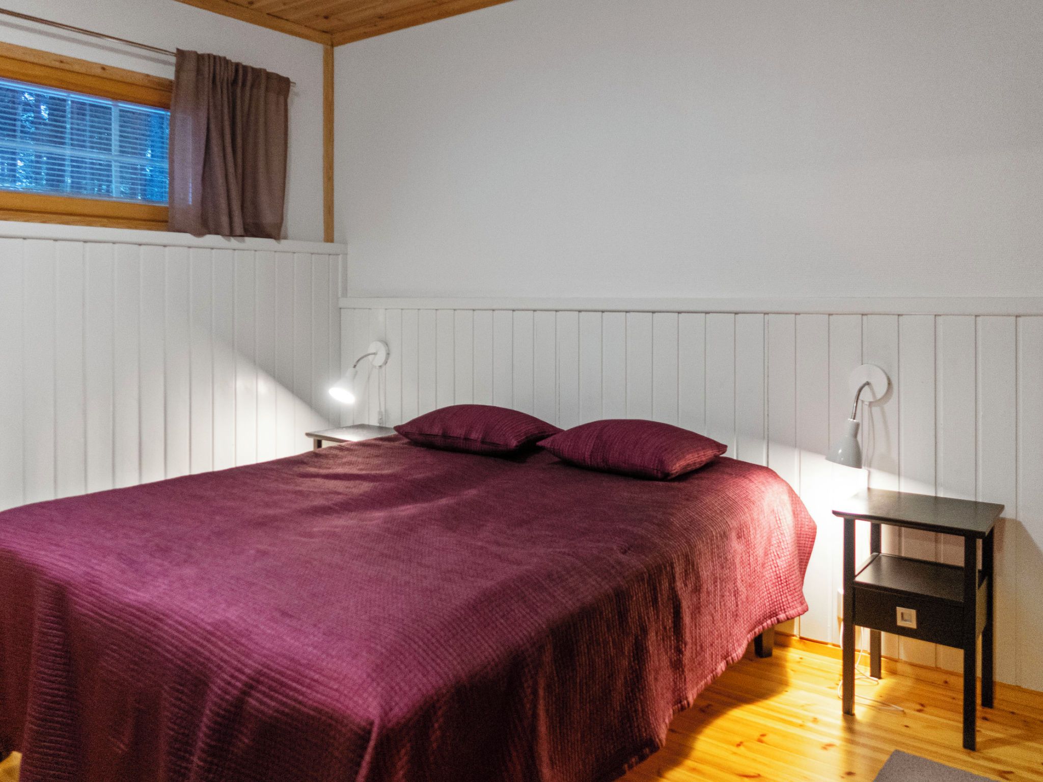 A bedroom with a bed, a nightstand, a window with curtains, and a wall-mounted lamp.