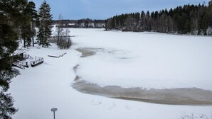 A snow-covered landscape with a frozen river, trees, and a small wooden structure.