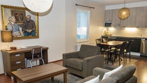 A modern living room with a sofa set, a wooden coffee table, and a desk with a lamp. A kitchen with a dining area is visible in the background.