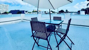 Una terraza en la azotea con un amplio paraguas, cuatro sillas de metal y una mesa de vidrio.