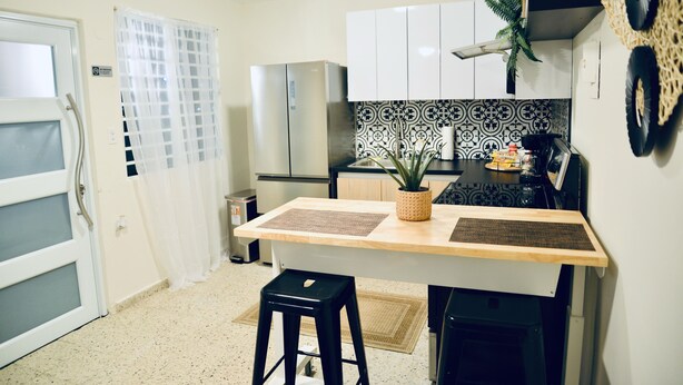 Una cocina compacta con encimera de madera, un taburete negro, un refrigerador y azulejos decorativos en la pared.