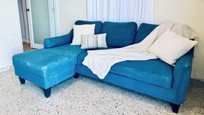 Un sofá azul con un plaid blanco y almohadas, una puerta y un suelo de baldosas.