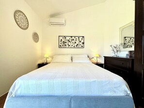 A neatly made bed with a white and blue striped duvet, two bedside tables with lamps, a wall-mounted air conditioner, and a framed floral artwork above the bed. - " Casa Luisa Trilocale " (Bari Sardo)