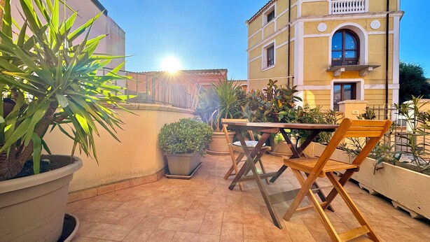 Eine Außenpatio mit Holztisch und Stühlen, umgeben von Topfpflanzen und einem Gebäude mit Balkon.