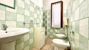 Un baño con lavamanos, inodoro y bidé, con azulejos en tonos verde y beige, una ventana y un espejo.
