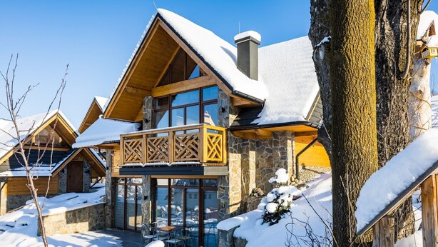 A chalet-style building with a wooden balcony and stone walls, surrounded by snow.