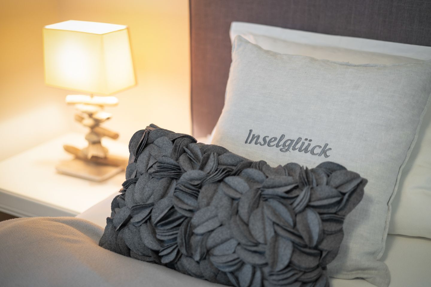A neatly made bed with a grey decorative pillow and a white pillow with the text "Inselglück" on it.