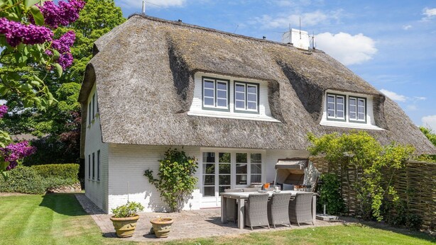 Une maison à toit de chaume, aux murs blancs, dotée d’une terrasse meublée de mobilier en osier et d’un jardin bien entretenu agrémenté de fleurs violettes.