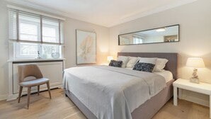 Une chambre à coucher comprenant un lit, une chaise, une table de chevet, une lampe, une fenêtre avec des stores et un tableau encadré accroché au mur.