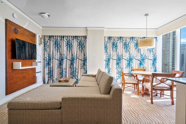 A modern living room with a sectional sofa, a flat-screen TV mounted on a wooden panel, a dining area with a table and chairs, and large windows with blue leaf-patterned curtains.