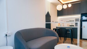 A modern living room with a grey sofa, a small white table, and a kitchen area with a bar counter and stools. - iSovo-iCity#3slep#Studio#netflix (Shah Alam)