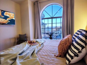 Un dormitorio con un gran ventanal arqueado, una cama con almohada a rayas y un cuadro en la pared.