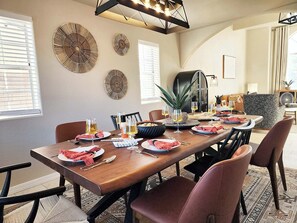 A dining area with a long wooden table set for a meal, surrounded by chairs, with decorative wall art and a modern light fixture above.
