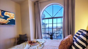 A bedroom with a large arched window, a bed with a quilt, pillows, a bedside table, and a wall-mounted TV.