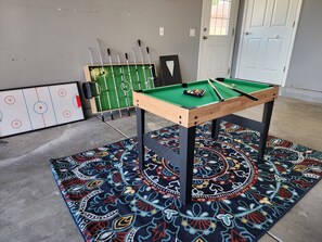 Una mesa de billar con un tapete estampado, una mesa de fútbol de mesa y una pista de hockey en una habitación con una puerta y una ventana.