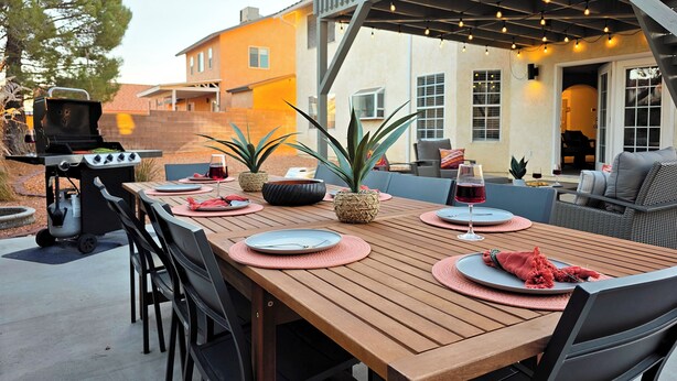 An outdoor patio with a wooden table set for dining, a grill, and a seating area with cushions.