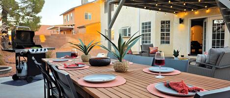 Un patio al aire libre con una mesa de madera dispuesta para comer, una parrilla y una zona de estar con cojines.