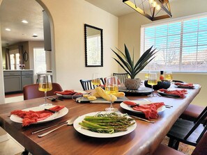 A dining area with a long table set for a meal, featuring plates of asparagus and bread, glasses of wine, and a potted plant.