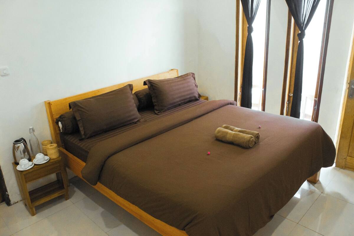 A bed with a brown bedspread, pillows, and a rolled towel. There is a small wooden nightstand with a kettle, cups, and a basket.