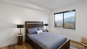 A modern bedroom with a wooden bed, two bedside lamps, a large window with a mountain view, and hardwood flooring. - Stunning Penthouse Breathtaking Water Views! 302 (Juneau)