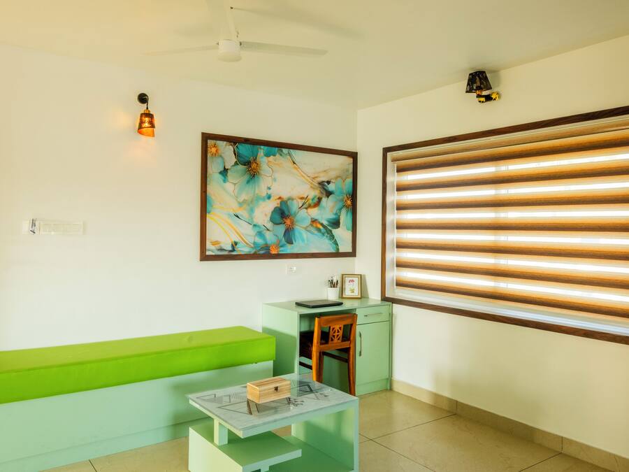 Una habitación con un banco verde, una mesita con una caja, un escritorio con una silla, un cuadro, un ventilador de techo y una ventana con persianas.