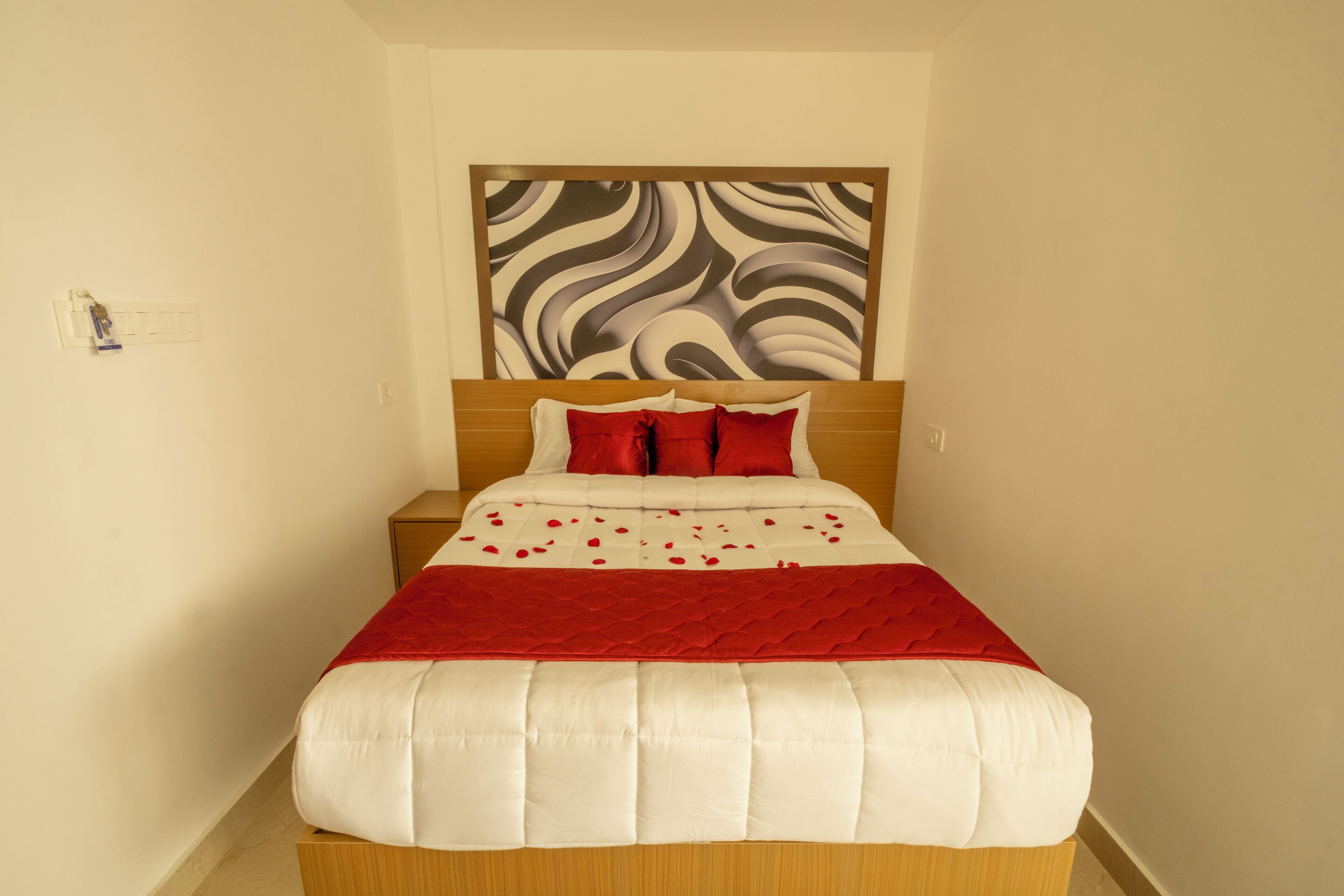 A neatly made bed with a red and white bedspread, a wooden headboard, and a wall with a large abstract art piece.