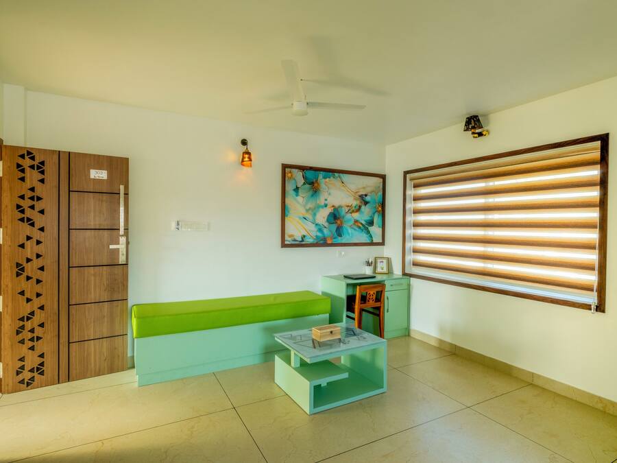 A room with a green sofa, a small table, a ceiling fan, a painting, and a window with blinds.