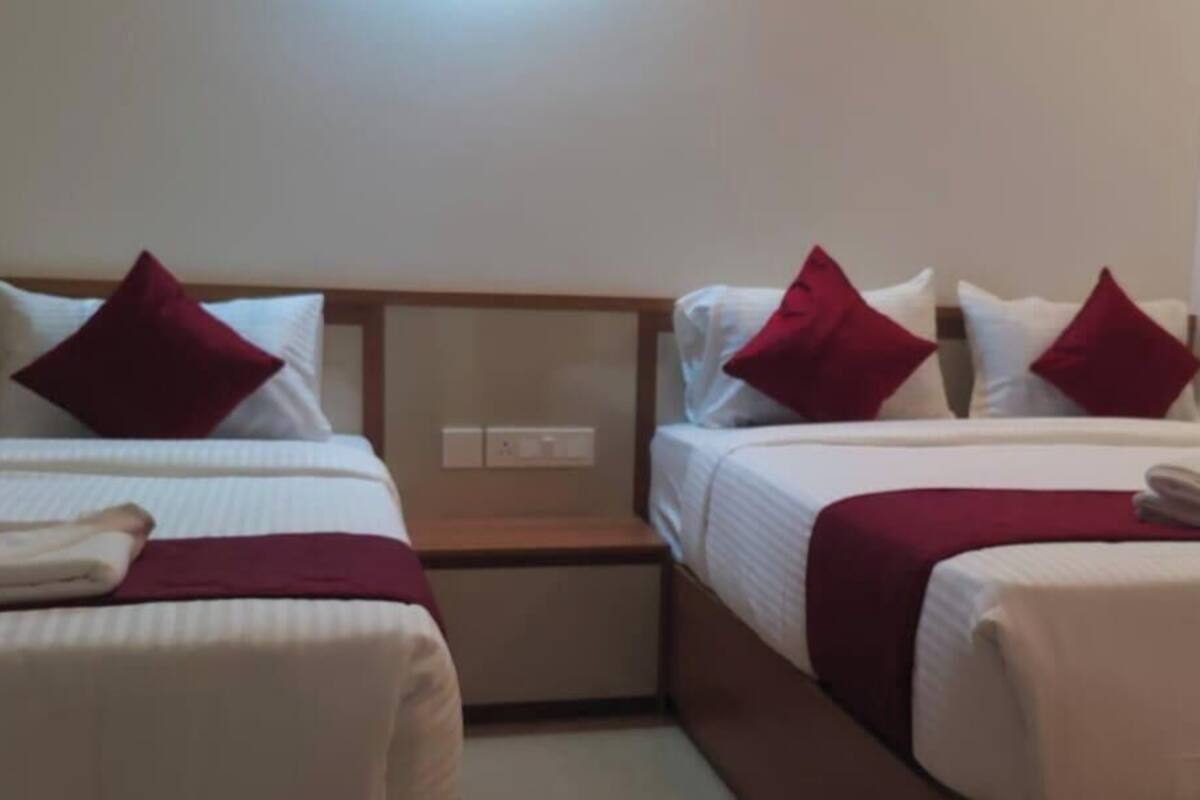 A hotel room with two single beds, each with a red pillow and white bedding. There is a wooden headboard, a nightstand with a lamp, and a folded towel on the bed.