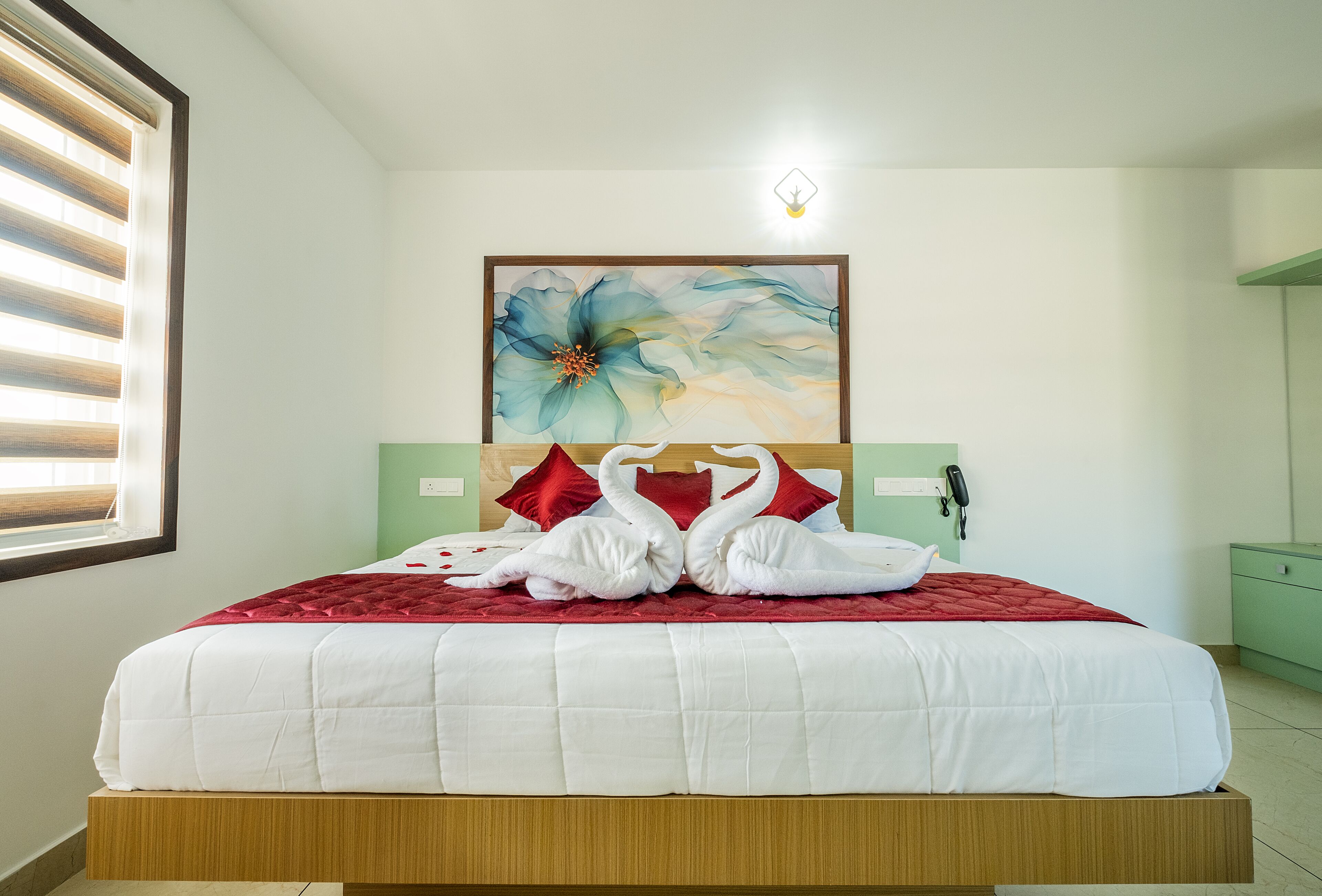 A neatly made bed with a red blanket and two white swan-shaped pillows, a large floral painting on the wall, and a bedside telephone.