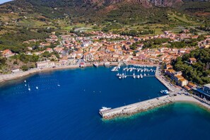 Vista aérea de un pueblo costero con un puerto deportivo, edificios residenciales y terreno montañoso al fondo.