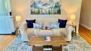 En stue med en beige sofa, to blå pude med blomstermønster, et træbord med en bog og et lys, samt et stort bjerglandskabsmaleri over sofaen.