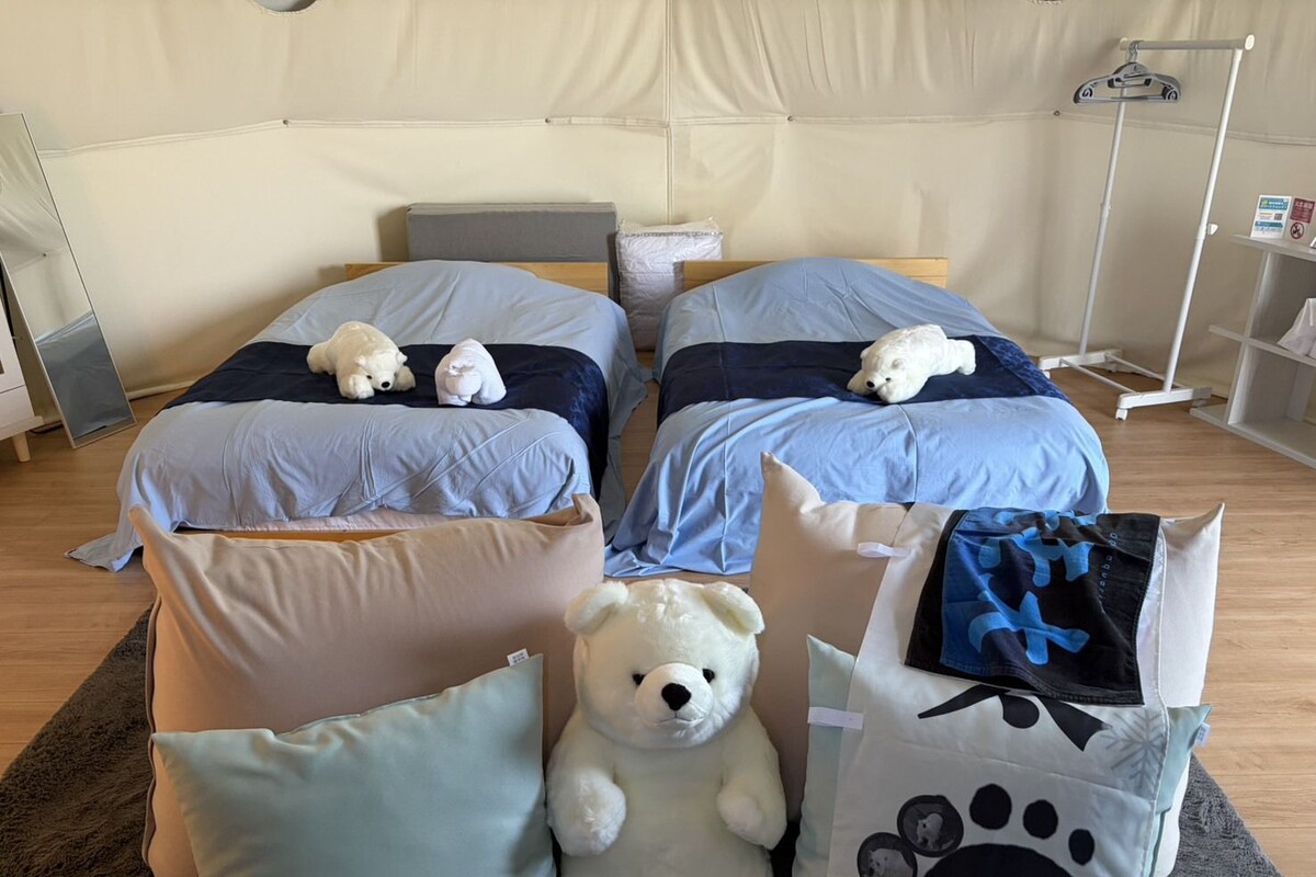 A room with two beds, each with a teddy bear, a large white teddy bear on the floor, and a bag with a paw print design.