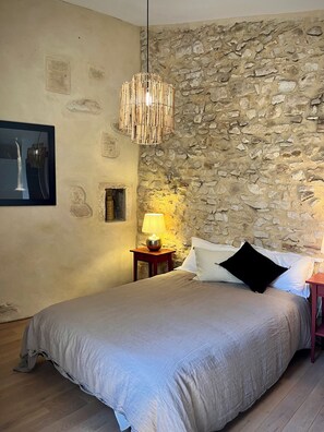 Une chambre avec un mur en pierre, un lit recouvert d’une couverture grise, une table de chevet en bois avec une lampe et un luminaire suspendu.