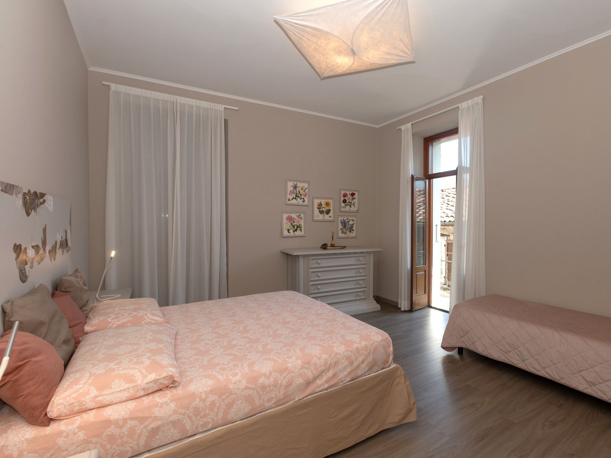 Une chambre avec un grand lit, une commode et une petite table. Il y a des rideaux aux fenêtres et une porte qui donne sur l’extérieur.