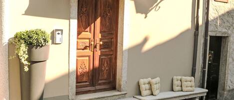 A building entrance with a wooden door, a planter with greenery, and a bench with cushions.