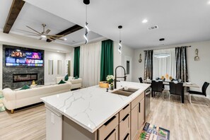 A modern living room with a large TV, a sofa, a dining area, and a kitchen with a marble island.