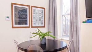 A round table with a black top surrounded by four clear chairs, a potted plant in the center, and two framed artworks on the wall. - Cozy Oasis | Near South End (Boston)