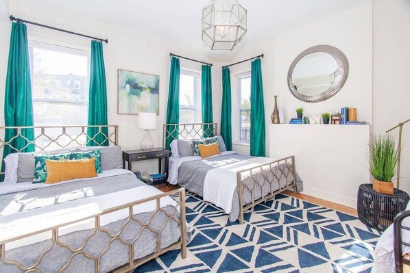 A bedroom with two beds, a geometric patterned rug, a bedside table with a lamp, a mirror, and a shelf with books and plants. - Expansive Essential Triplex | near T (Boston)