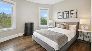 Un dormitorio con una cama grande, dos ventanas, un radiador y tres cuadros colgados en la pared.