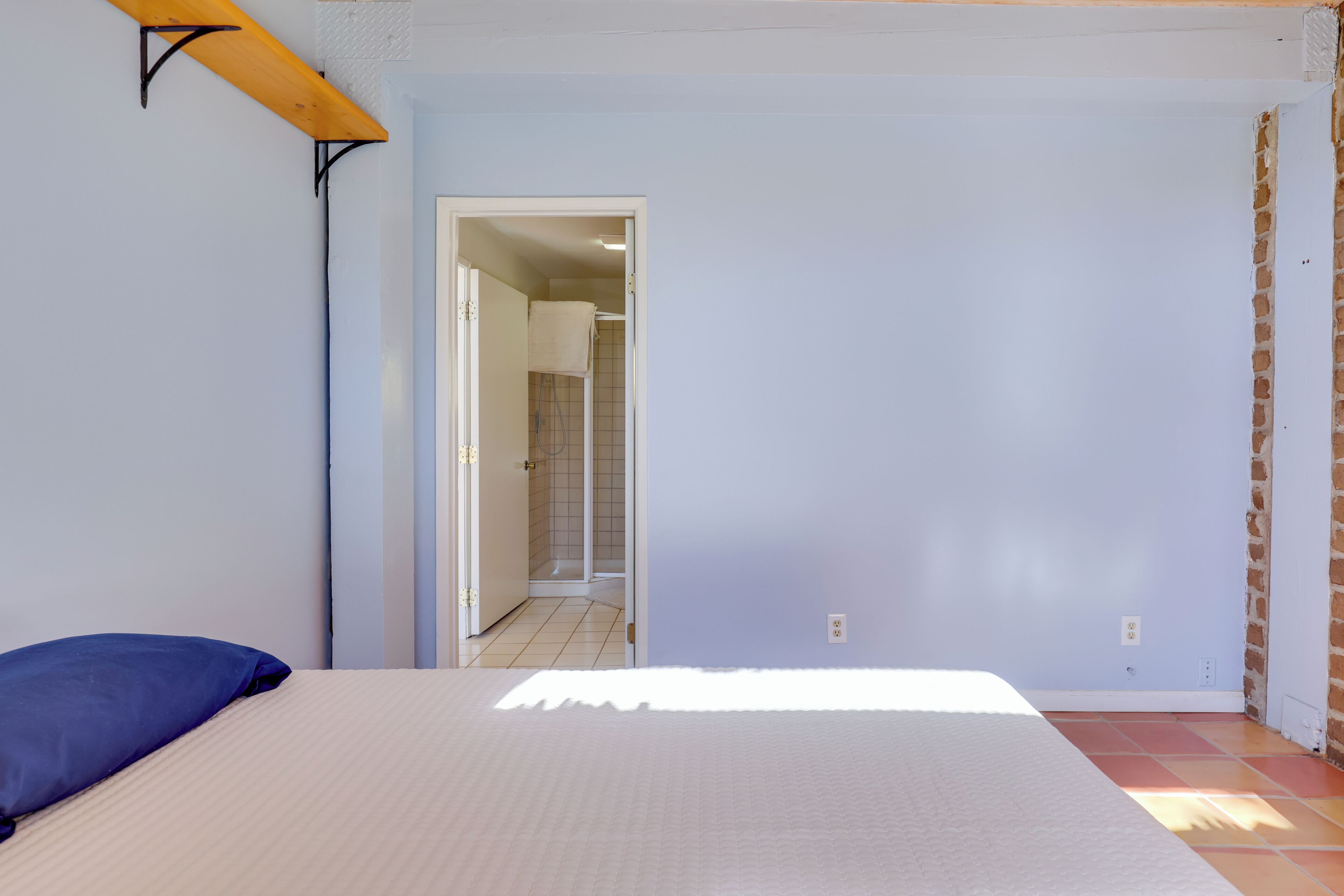 A bedroom with a bed, a blue pillow, a wooden shelf, and a door leading to a bathroom.