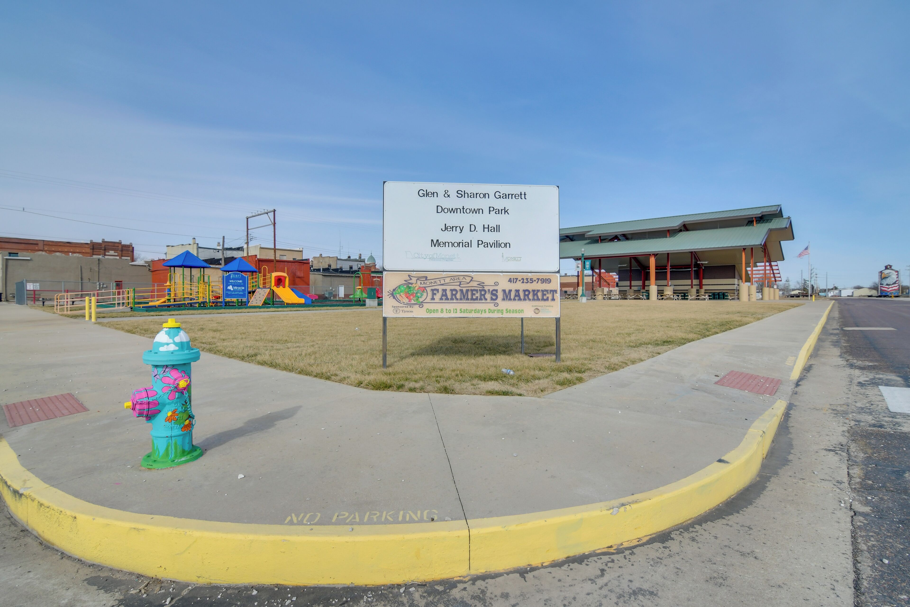 A fire hydrant with a colorful design, a sign for Glen & Sharon Garrett Downtown Park, Jerry D. Hall Memorial Pavilion, and a Farmer's Market sign.