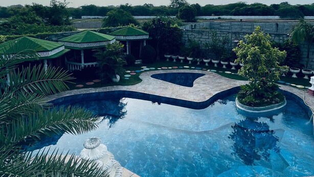 A large, curved swimming pool with a stone border, surrounded by lush greenery and a gazebo. - Sparrow Farms And Resort (Mohanlalganj)