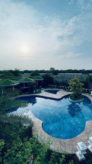 A large, curved swimming pool with a stone border, surrounded by lush greenery and a gazebo. - Sparrow Farms And Resort (Mohanlalganj)