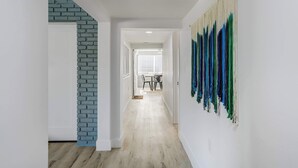 A hallway with a staircase, a dining area in the background, and a wall with a textured piece of art. - Riverside Luau! Tiki Hut, Fire Pit & Outdoor Fun (Bradenton)