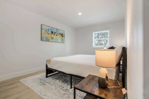 A bedroom with a bed, a bedside table with a lamp, a window, and a painting on the wall.