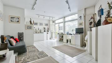 A spacious living area with a sofa, coffee table, and artwork on the walls. A kitchen area with a dining table and chairs is visible in the background.