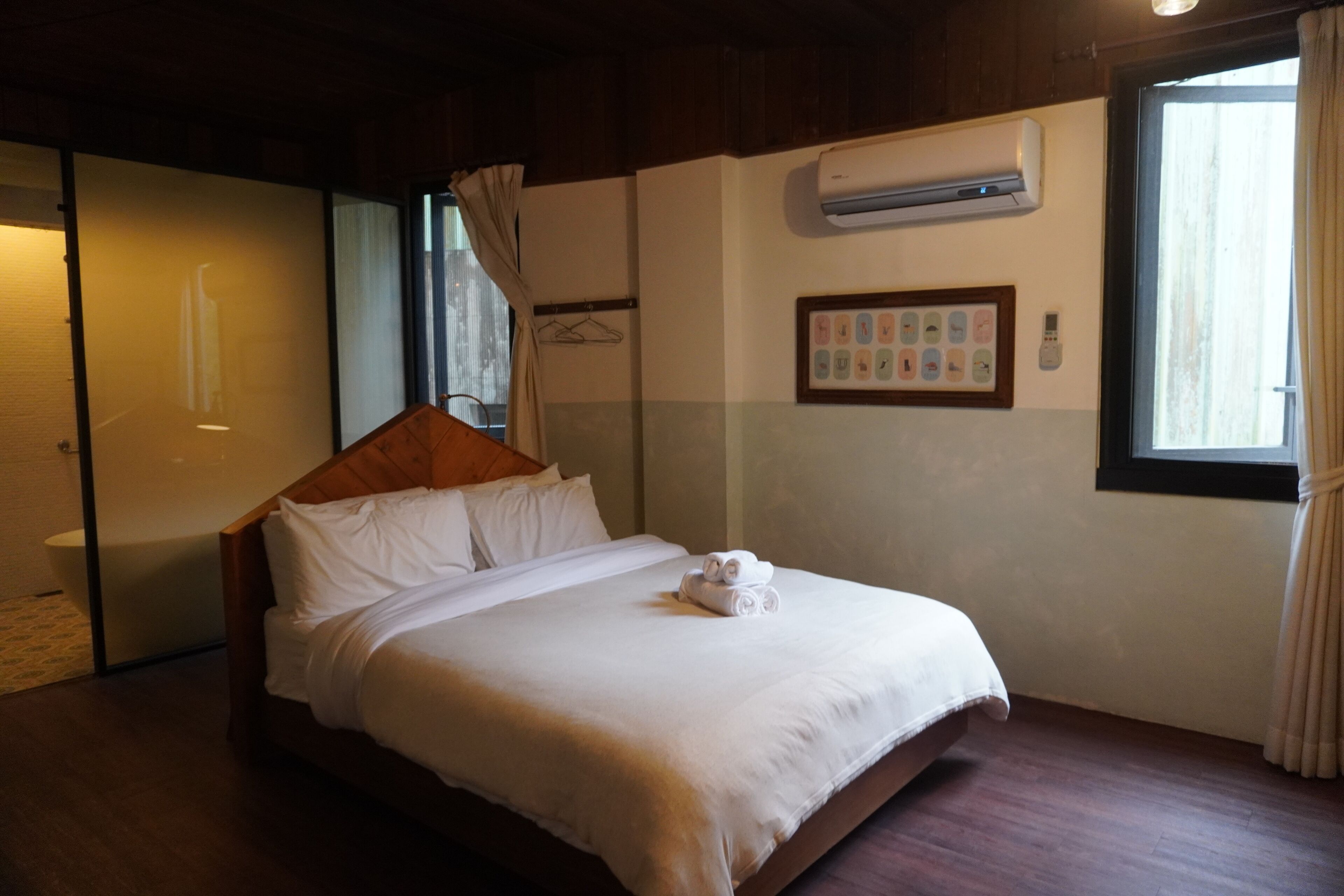 A neatly made bed with white linens and a rolled towel, a wooden headboard, a wall-mounted air conditioner, a framed picture of children's activities, and a window with curtains.