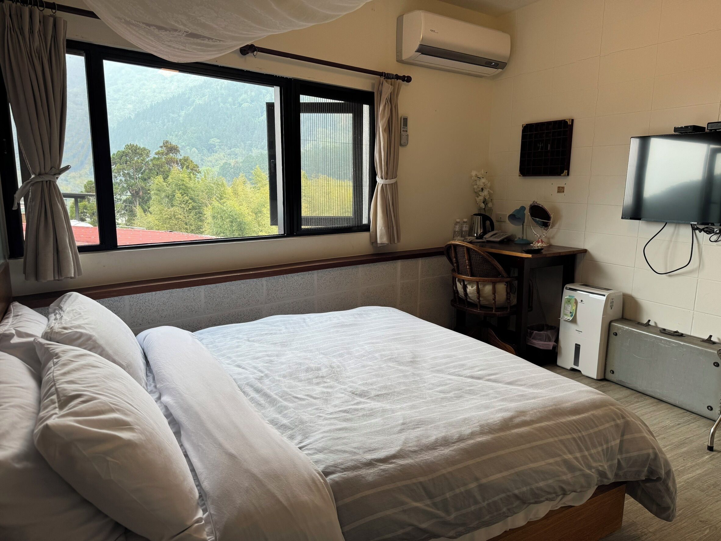 A hotel room with a bed, a desk, a chair, an air conditioner, a television, and a view of the outdoors.